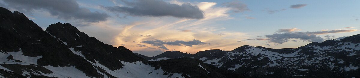 Vue des Alpes-Maritimes à la fin du printemps, avec les sommets encore enneigés illuminés par la lumière du coucher du soleil et le ciel traversé de nuages dorés. Archives APAM. Fin de la description de l’image.