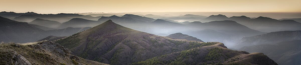 Vue panoramique de la Cima Gouron, avec la lumière dorée se fondant sur les crêtes montagneuses et les vallées plongées dans une légère brume. Archives APAM. Fin de la description de l’image.