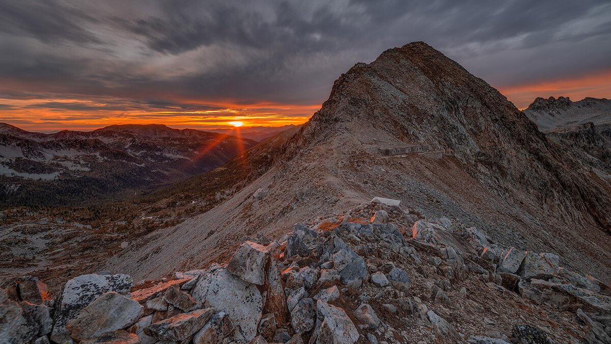 Vue du col transfrontalier de Fremamorta, avec le soleil illuminant de lumière orangée les roches et le sentier de crête. Photo de M. Lucotti. Fin de la description de l’image.