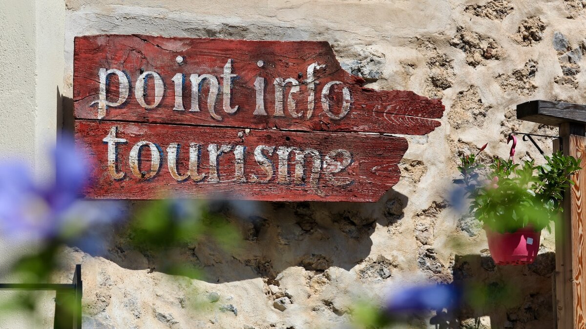 Panneau touristique en bois rouge portant l’inscription « point info tourisme », fixé sur un mur en pierre, avec des fleurs floues au premier plan. Fin de la description de l’image.